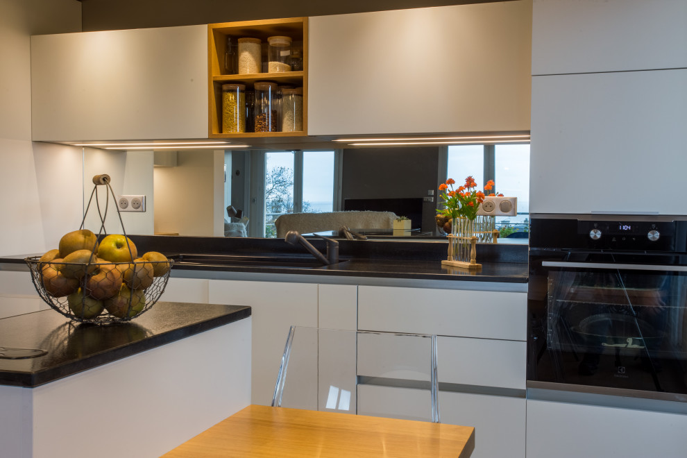 Mid-sized modern l-shaped open plan kitchen in Other with an integrated sink, beaded inset cabinets, white cabinets, granite benchtops, mirror splashback, stainless steel appliances, cement tiles, with island, multi-coloured floor and black benchtop.