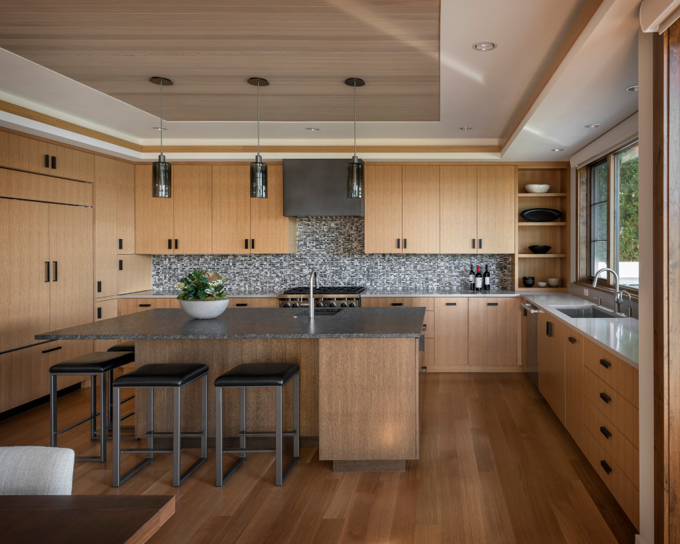 Large contemporary u-shaped eat-in kitchen in Seattle with an undermount sink, flat-panel cabinets, light wood cabinets, granite benchtops, grey splashback, mosaic tile splashback, panelled appliances, light hardwood floors, with island, black benchtop and wood.