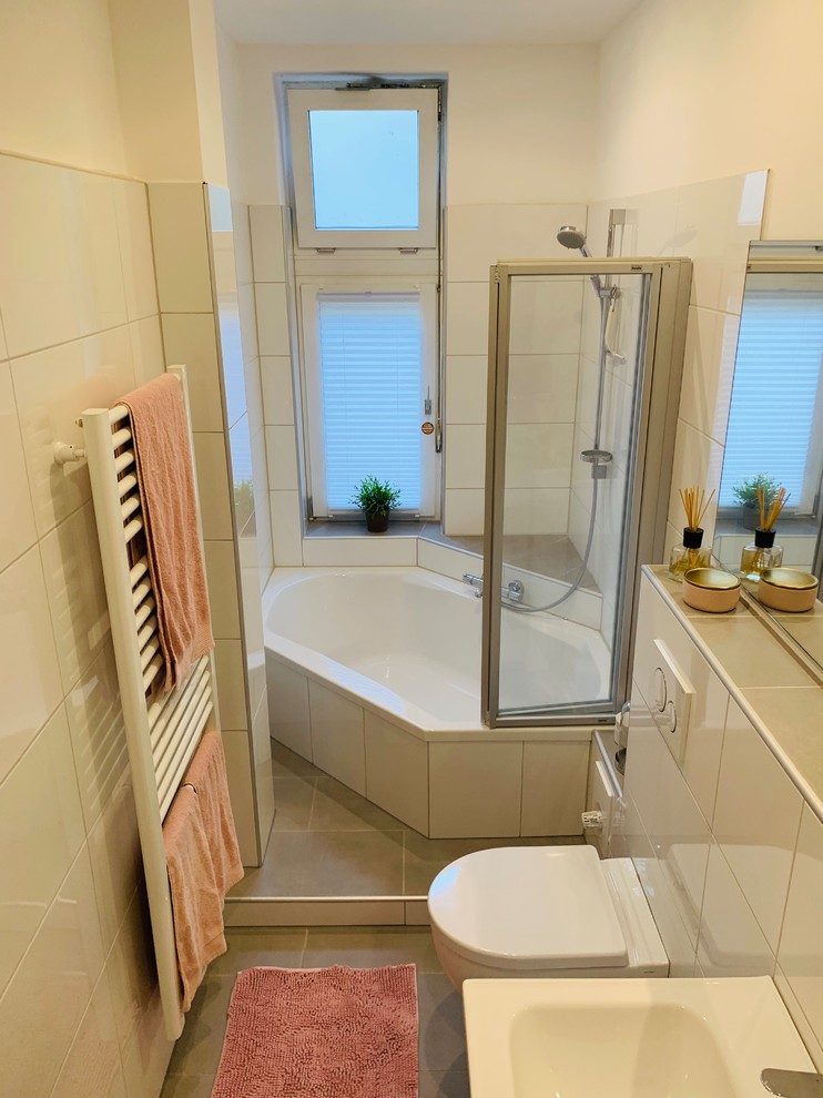This is an example of a mid-sized contemporary 3/4 bathroom in Hanover with white cabinets, a corner tub, a shower/bathtub combo, a wall-mount toilet, white tile, ceramic tile, white walls, ceramic floors, a wall-mount sink, solid surface benchtops, grey floor, a hinged shower door and white benchtops.