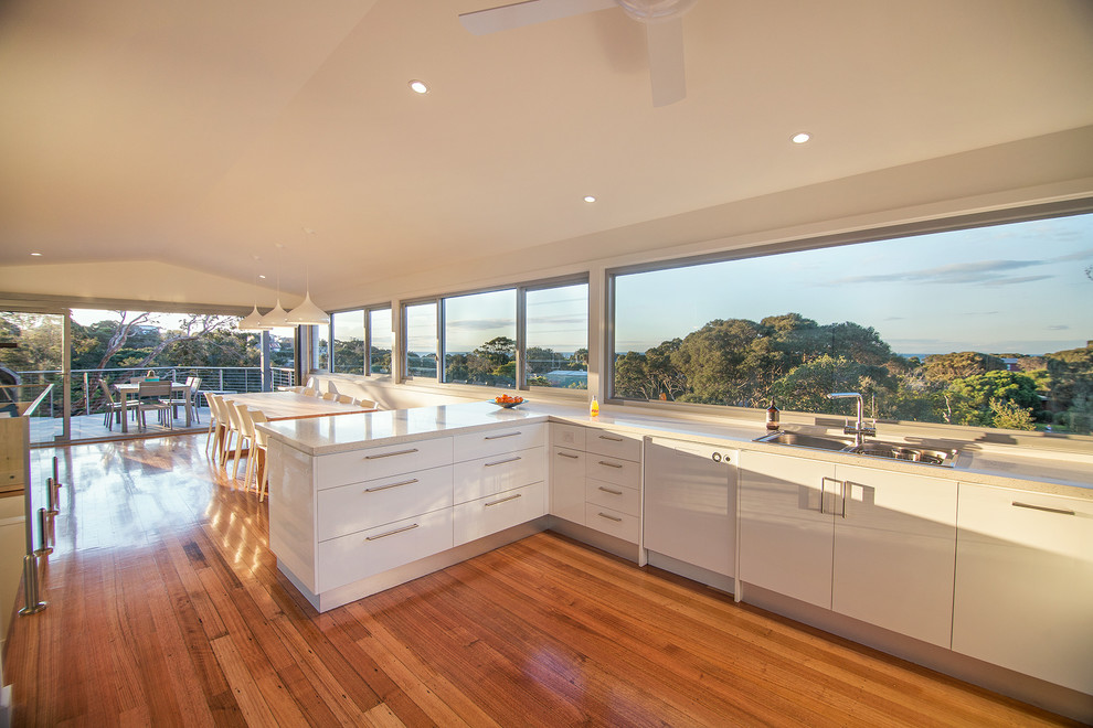 Photo of a large beach style u-shaped eat-in kitchen in Melbourne with a drop-in sink, white cabinets, quartz benchtops, glass sheet splashback and stainless steel appliances.