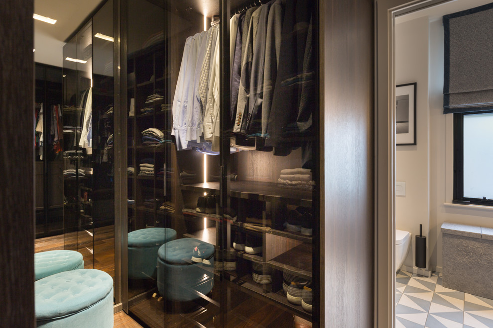 This is an example of a small contemporary gender-neutral walk-in wardrobe in Gloucestershire with glass-front cabinets, dark wood cabinets, dark hardwood floors and brown floor.
