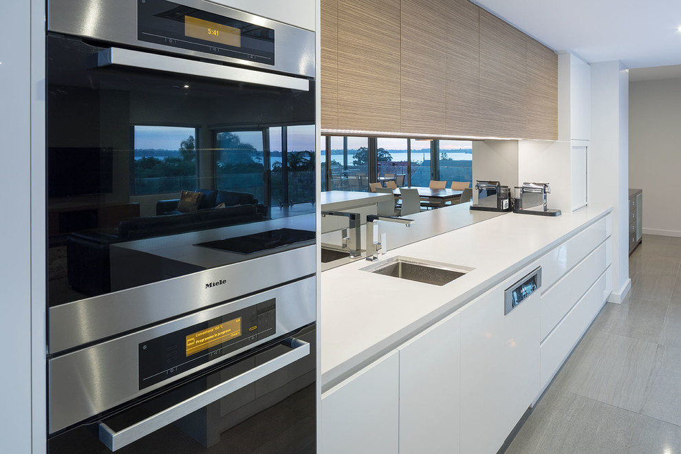 Design ideas for a large modern galley open plan kitchen in Perth with an undermount sink, flat-panel cabinets, white cabinets, solid surface benchtops, metallic splashback, mirror splashback, stainless steel appliances, ceramic floors, with island, grey floor and white benchtop.