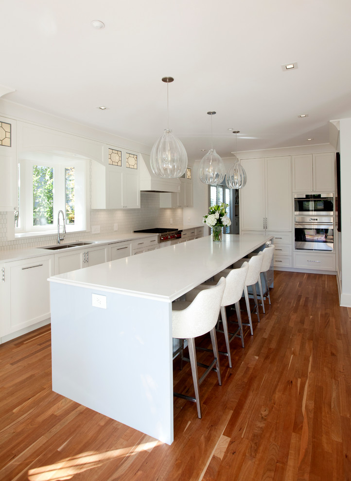 Photo of a mid-sized contemporary u-shaped open plan kitchen in Vancouver with a double-bowl sink, recessed-panel cabinets, white cabinets, solid surface benchtops, white splashback, ceramic splashback, stainless steel appliances, medium hardwood floors, with island, brown floor and white benchtop.
