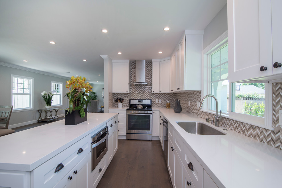 Photo of a mid-sized traditional l-shaped eat-in kitchen in Los Angeles with an undermount sink, shaker cabinets, white cabinets, solid surface benchtops, multi-coloured splashback, stainless steel appliances, dark hardwood floors, with island, brown floor and white benchtop.