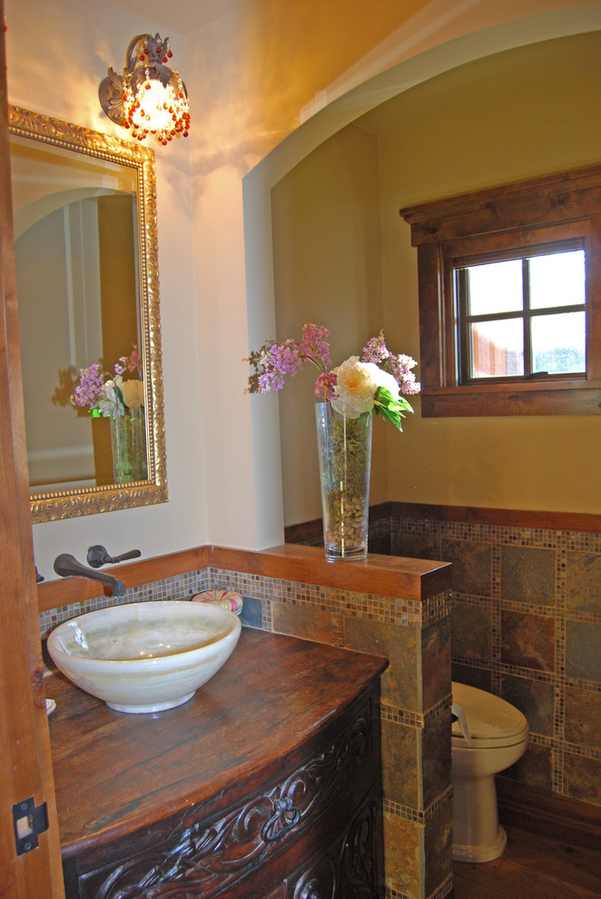 Design ideas for a mid-sized country powder room in Minneapolis with a vessel sink, furniture-like cabinets, dark wood cabinets, wood benchtops, multi-coloured tile, porcelain tile, beige walls and brown benchtops.