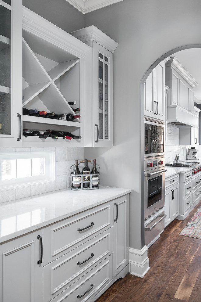 Large transitional l-shaped eat-in kitchen in Chicago with an undermount sink, white cabinets, quartz benchtops, white splashback, ceramic splashback, stainless steel appliances, medium hardwood floors, with island, brown floor and white benchtop.