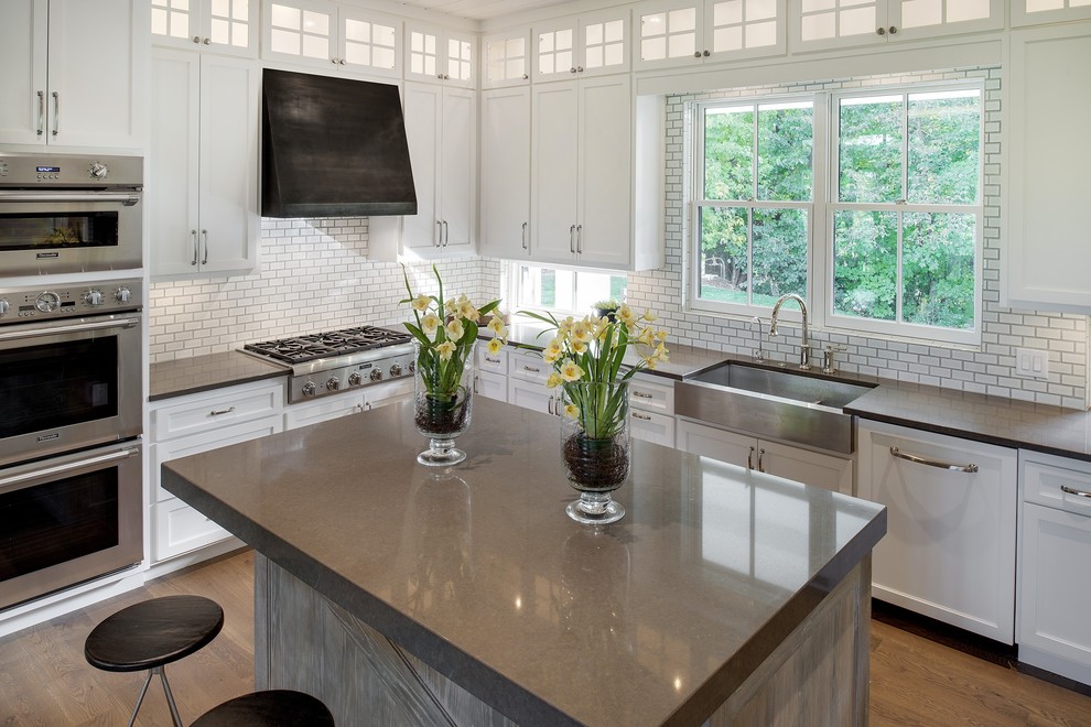 This is an example of a mid-sized country u-shaped open plan kitchen in Minneapolis with a farmhouse sink, shaker cabinets, white cabinets, white splashback, subway tile splashback, stainless steel appliances, light hardwood floors, with island and brown floor.