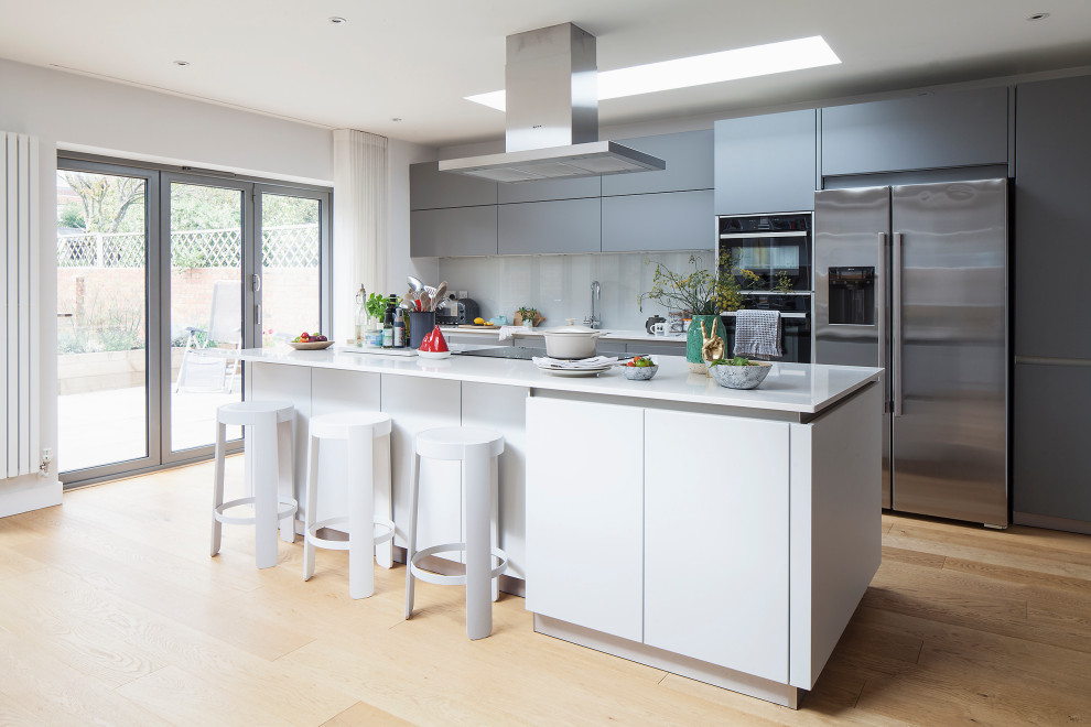 Photo of a large scandinavian single-wall open plan kitchen in Sussex with an integrated sink, flat-panel cabinets, grey cabinets, solid surface benchtops, green splashback, glass sheet splashback, stainless steel appliances, medium hardwood floors, with island, brown floor and white benchtop.