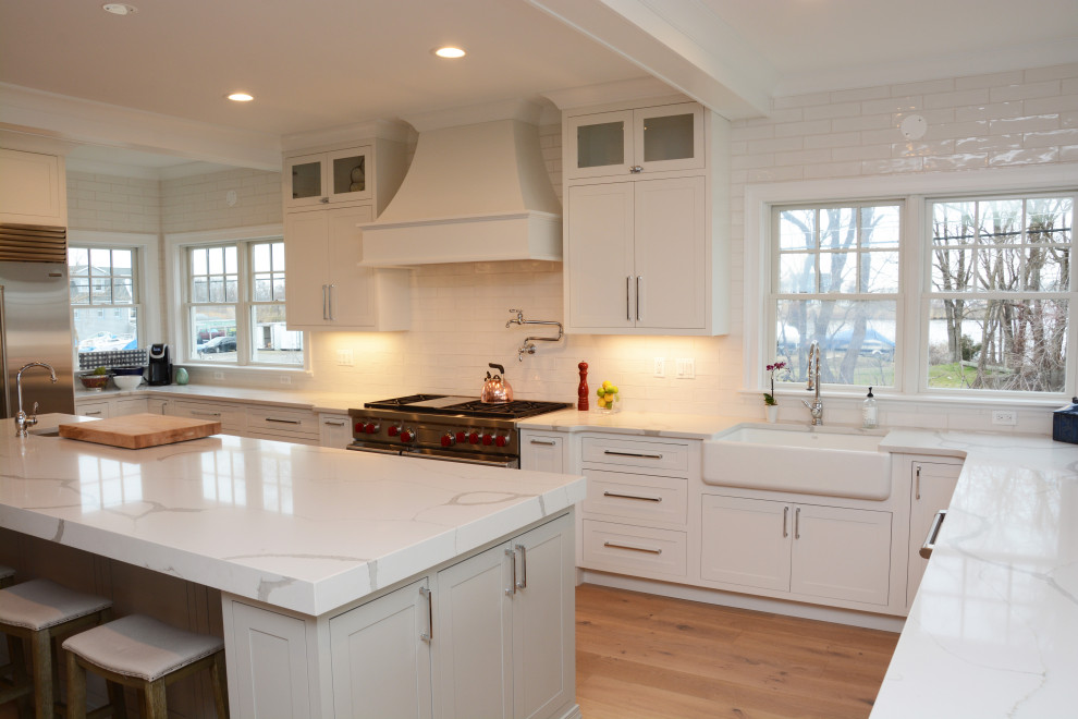Klassisk inredning av ett stort vit vitt kök, med en rustik diskho, luckor med profilerade fronter, vita skåp, bänkskiva i kvarts, vitt stänkskydd, rostfria vitvaror, en köksö och brunt golv