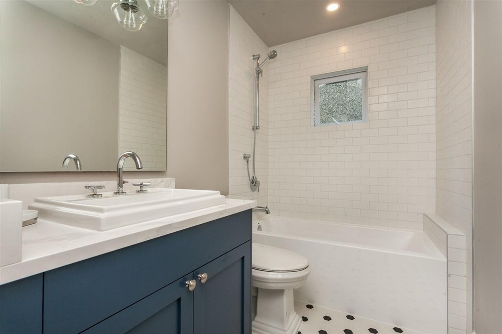 Photo of a mid-sized beach style 3/4 bathroom in Vancouver with shaker cabinets, blue cabinets, an alcove tub, a two-piece toilet, white tile, ceramic tile, beige walls, ceramic floors, a drop-in sink, engineered quartz benchtops, white floor and white benchtops.