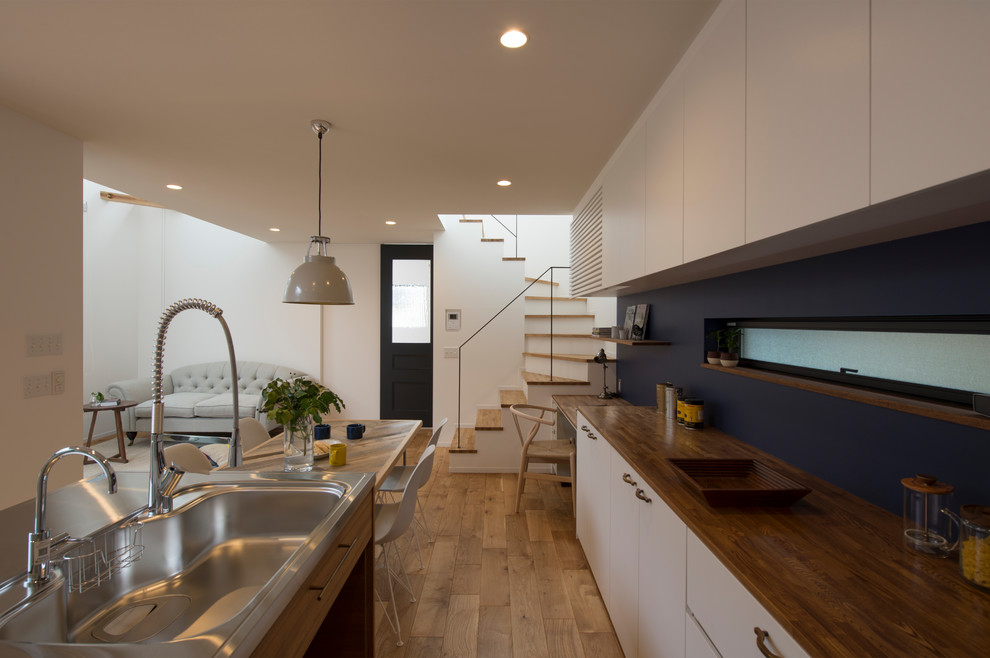Photo of a scandinavian single-wall open plan kitchen in Other with an integrated sink, flat-panel cabinets, white cabinets, stainless steel benchtops, light hardwood floors, a peninsula, beige floor and brown benchtop.