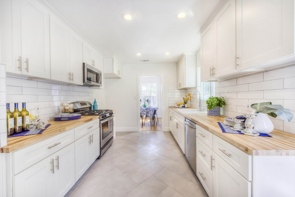 Beach style galley separate kitchen in Los Angeles with a drop-in sink, shaker cabinets, white cabinets, wood benchtops, white splashback, ceramic splashback, stainless steel appliances, no island, grey floor and beige benchtop.