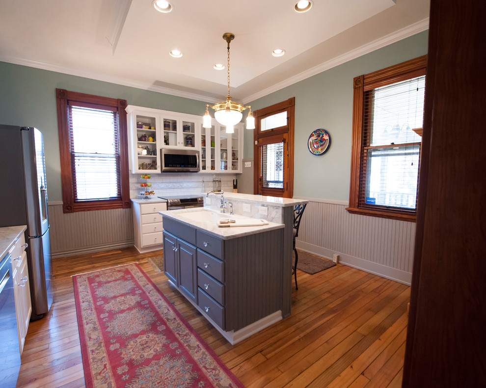Mid-sized eclectic u-shaped light wood floor eat-in kitchen photo in Columbus with a drop-in sink, raised-panel cabinets, white cabinets, marble countertops, white backsplash, stone tile backsplash, stainless steel appliances and an island
