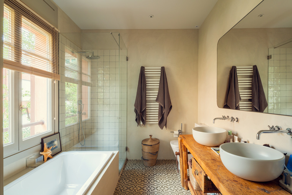 This is an example of a mediterranean master bathroom in Barcelona with a vessel sink, a hinged shower door, medium wood cabinets, a drop-in tub, a corner shower, a one-piece toilet, white tile, ceramic tile, beige walls, wood benchtops, multi-coloured floor, brown benchtops and flat-panel cabinets.