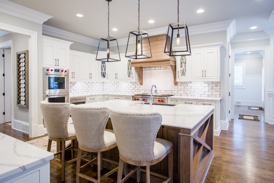 This is an example of a large transitional u-shaped eat-in kitchen in Atlanta with a farmhouse sink, raised-panel cabinets, white cabinets, quartz benchtops, white splashback, brick splashback, stainless steel appliances, medium hardwood floors, with island, brown floor and white benchtop.