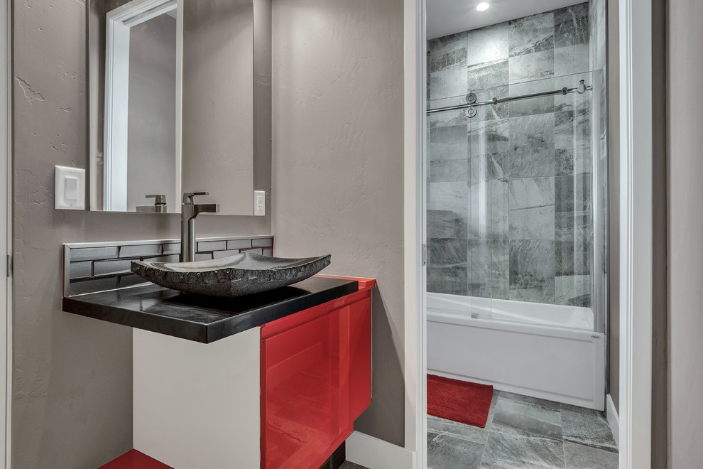 Inspiration for a mid-sized contemporary bathroom in Denver with flat-panel cabinets, red cabinets, a drop-in tub, a shower/bathtub combo, black tile, porcelain tile, grey walls, porcelain floors, a vessel sink, quartzite benchtops, grey floor, a sliding shower screen and black benchtops.