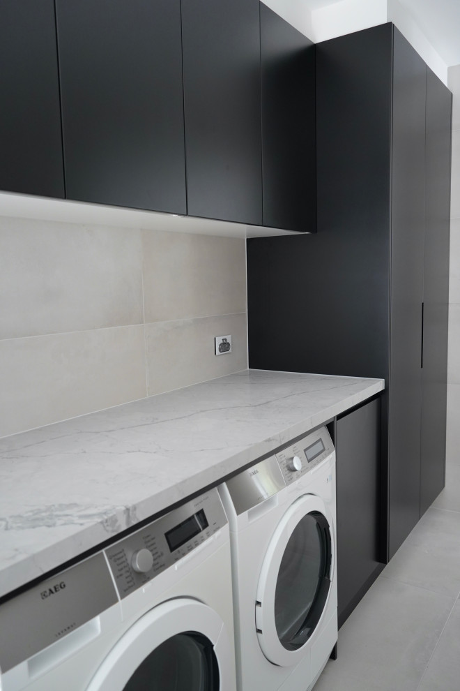 Mid-sized modern single-wall dedicated laundry room in Sydney with a drop-in sink, flat-panel cabinets, black cabinets, quartz benchtops, grey splashback, cement tile splashback, white walls, a side-by-side washer and dryer and grey benchtop.