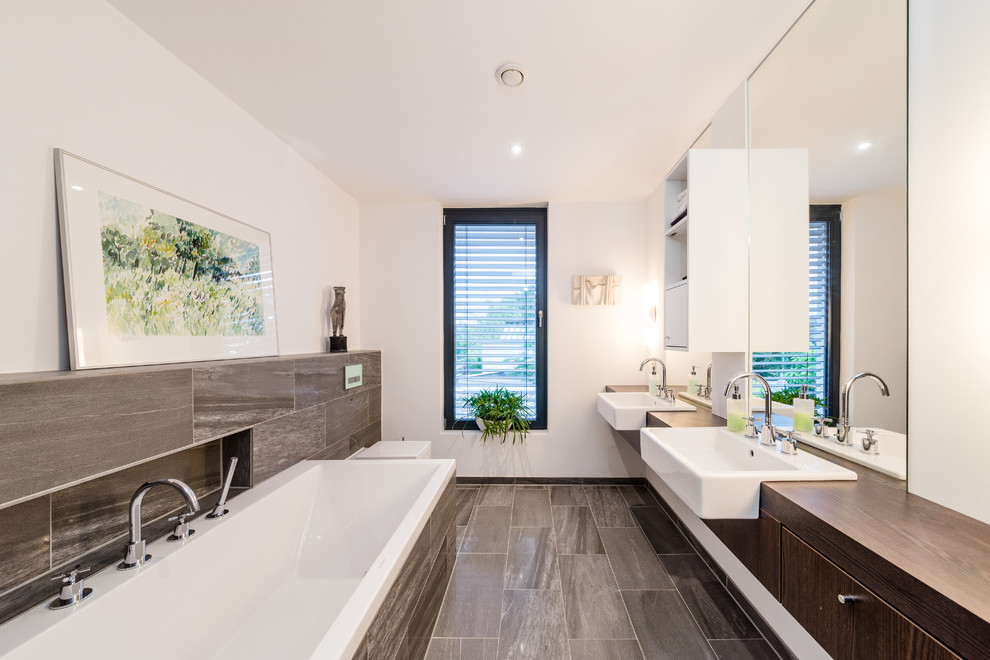 Großes Modernes Badezimmer En Suite mit Aufsatzwaschbecken, flächenbündigen Schrankfronten, dunklen Holzschränken, Waschtisch aus Holz, Einbaubadewanne, braunen Fliesen, weißer Wandfarbe, Keramikboden, Steinfliesen, Wandtoilette und brauner Waschtischplatte in Frankfurt am Main