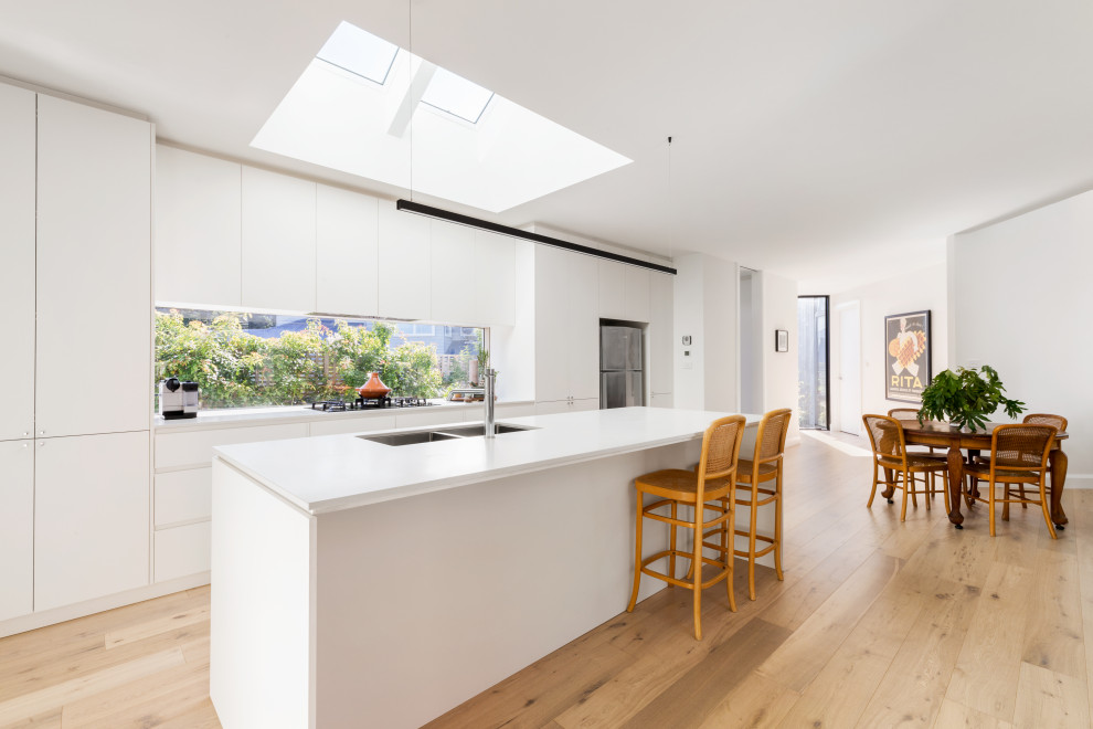Mid-sized scandinavian galley open plan kitchen in Melbourne with an undermount sink, flat-panel cabinets, white cabinets, quartz benchtops, window splashback, stainless steel appliances, light hardwood floors, with island, beige floor and white benchtop.