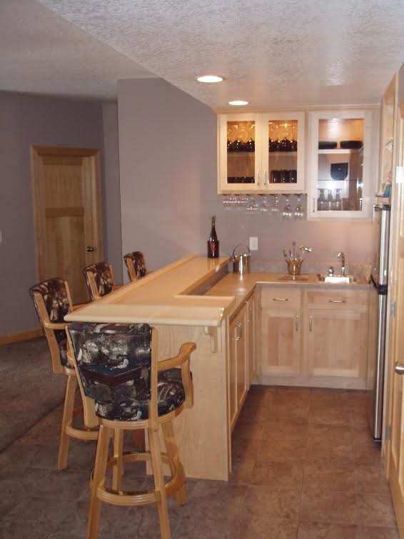 This is an example of a small traditional l-shaped seated home bar in Minneapolis with glass-front cabinets, light wood cabinets, wood benchtops, travertine floors and beige floor.