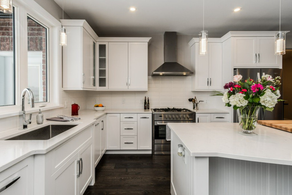 Mid-sized transitional l-shaped dark wood floor and brown floor open concept kitchen photo in Ottawa with an undermount sink, shaker cabinets, white cabinets, quartz countertops, white backsplash, subway tile backsplash, stainless steel appliances, an island and white countertops