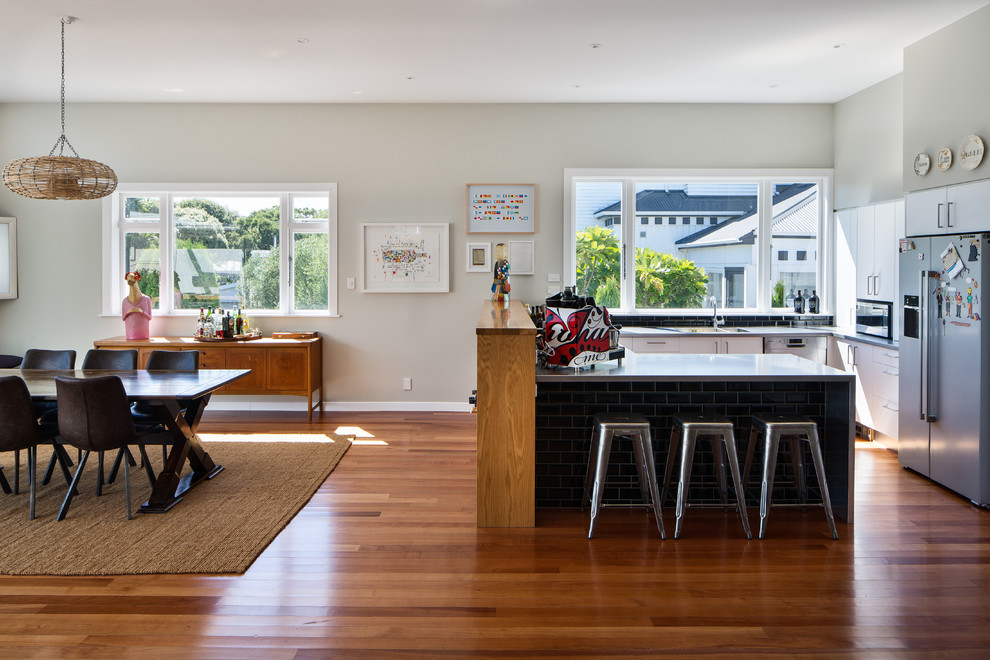 This is an example of a contemporary u-shaped open plan kitchen in Wellington with a double-bowl sink, flat-panel cabinets, white cabinets, stainless steel benchtops, black splashback, subway tile splashback, stainless steel appliances, medium hardwood floors, a peninsula, brown floor and grey benchtop.
