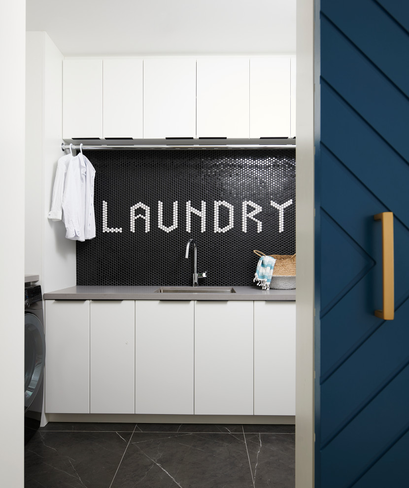 Photo of a mid-sized contemporary l-shaped dedicated laundry room in Toronto with an undermount sink, flat-panel cabinets, white cabinets, quartz benchtops, black splashback, mosaic tile splashback, white walls, porcelain floors, a side-by-side washer and dryer, black floor and grey benchtop.