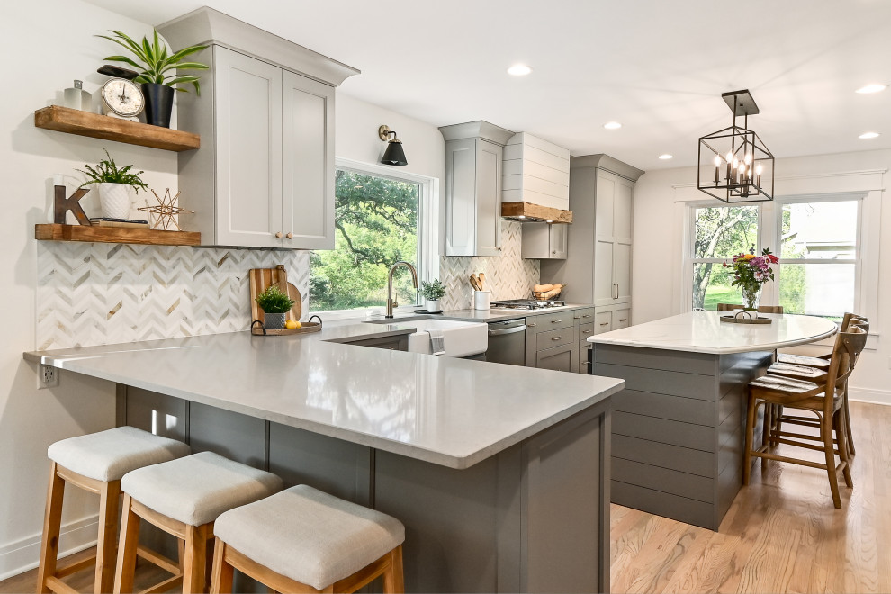 This is an example of an expansive country l-shaped eat-in kitchen in Milwaukee with a farmhouse sink, shaker cabinets, grey cabinets, quartz benchtops, white splashback, marble splashback, stainless steel appliances, medium hardwood floors, with island, brown floor and grey benchtop.