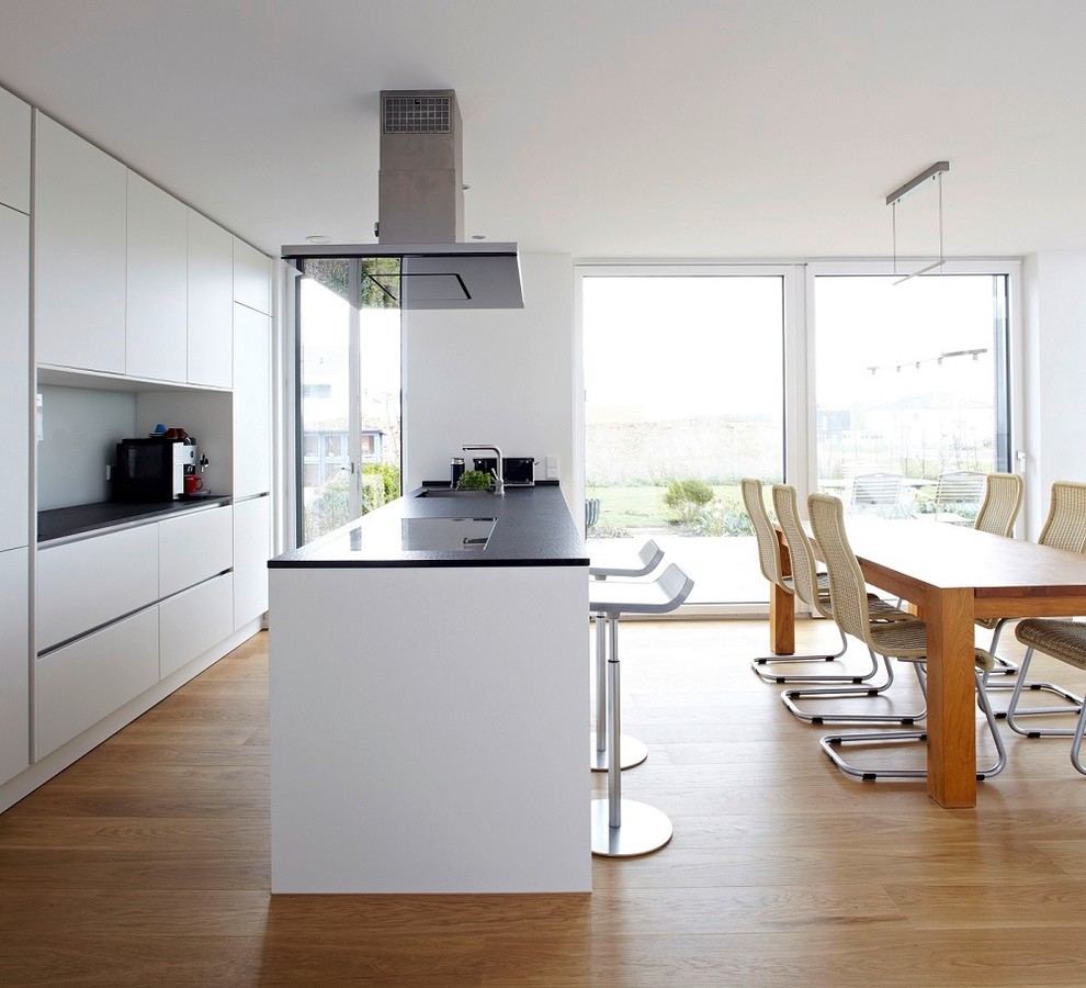 This is an example of a mid-sized modern galley open plan kitchen in Hanover with a single-bowl sink, flat-panel cabinets, white cabinets, white splashback, glass sheet splashback, stainless steel appliances, medium hardwood floors and with island.
