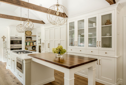 Airy farmhouse kitchen with white shaker RTA cabinets, glass-front pantry wall, vaulted ceiling with wood beams, globe pendants, and an island cooktop with a wood table extension.