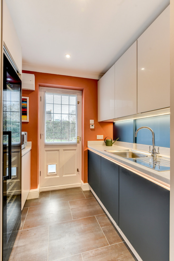 Photo of a large modern u-shaped eat-in kitchen in Sussex with a drop-in sink, flat-panel cabinets, blue cabinets, solid surface benchtops, blue splashback, glass sheet splashback, panelled appliances, vinyl floors, no island, brown floor and white benchtop.