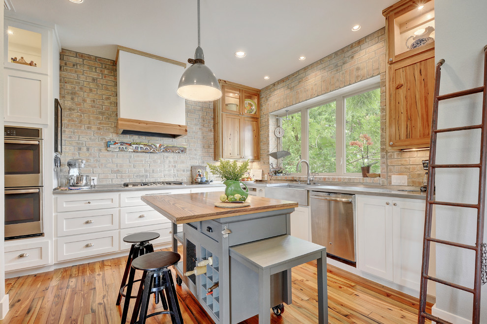 Photo of an industrial l-shaped kitchen in Austin with a farmhouse sink, shaker cabinets, white cabinets, brick splashback, stainless steel appliances, medium hardwood floors and with island.