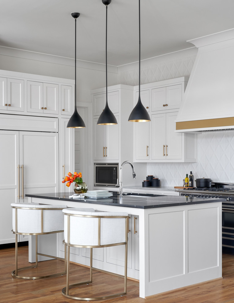 Example of a mid-sized transitional l-shaped medium tone wood floor and brown floor kitchen design in Dallas with an undermount sink, white cabinets, white backsplash, ceramic backsplash, an island, shaker cabinets and black countertops