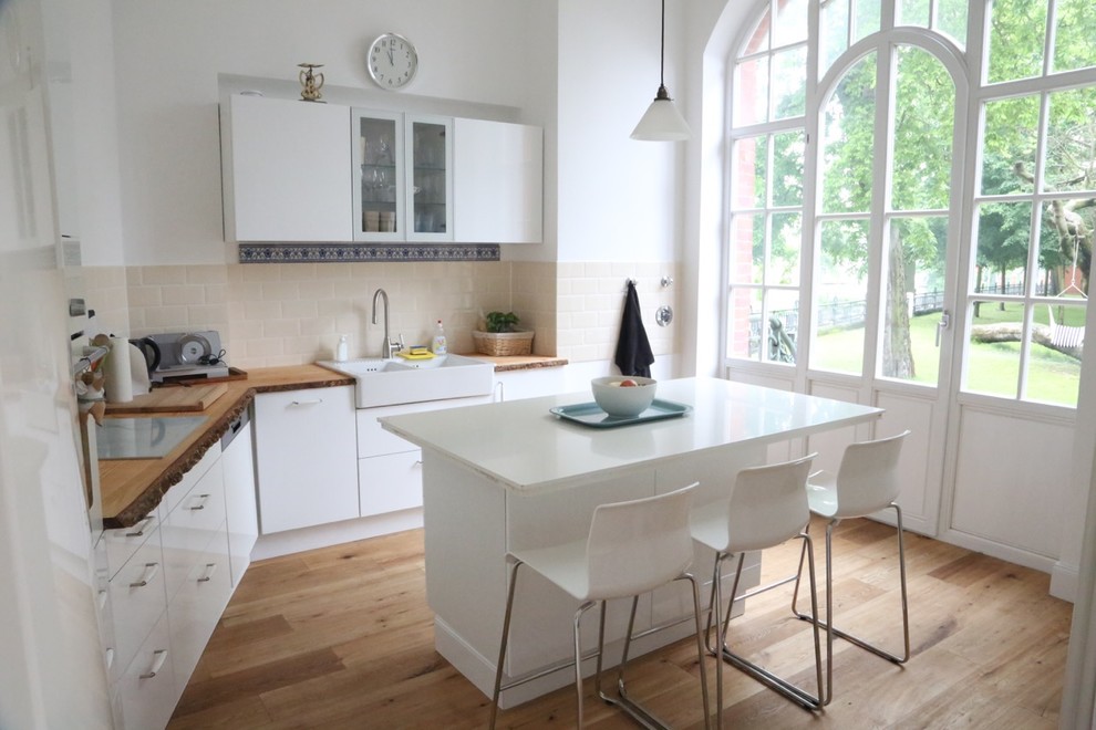 Photo of a mid-sized traditional l-shaped separate kitchen in Berlin with a farmhouse sink, flat-panel cabinets, white cabinets, wood benchtops, beige splashback, subway tile splashback, white appliances, medium hardwood floors and with island.