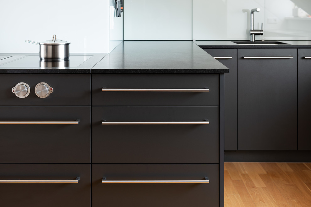 This is an example of a mid-sized contemporary eat-in kitchen in Munich with flat-panel cabinets, black cabinets, granite benchtops, white splashback, glass sheet splashback, black appliances, medium hardwood floors, no island, brown floor and black benchtop.