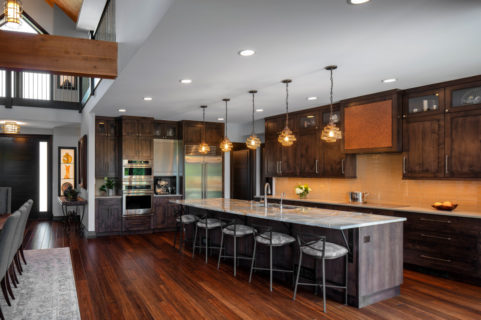 This is an example of a country l-shaped open plan kitchen in Chicago with an undermount sink, raised-panel cabinets, dark wood cabinets, marble benchtops, orange splashback, glass tile splashback, stainless steel appliances, dark hardwood floors, with island, brown floor and grey benchtop.