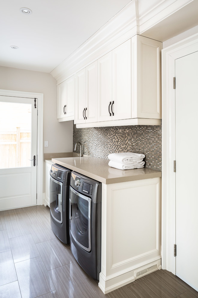 This is an example of a large traditional galley utility room in Toronto with a single-bowl sink, recessed-panel cabinets, white cabinets, quartz benchtops, beige walls, ceramic floors, a side-by-side washer and dryer and brown floor.