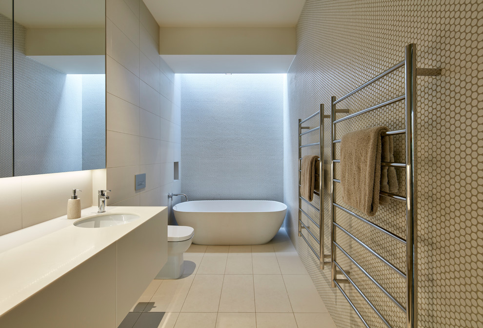 Design ideas for a mid-sized contemporary kids bathroom in Melbourne with flat-panel cabinets, white cabinets, a freestanding tub, a one-piece toilet, beige tile, mosaic tile, beige walls, porcelain floors, an undermount sink, solid surface benchtops, beige floor and white benchtops.