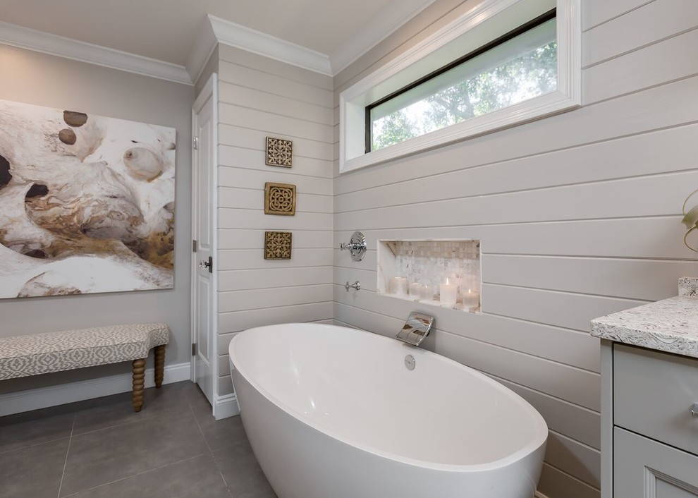 This is an example of a mid-sized beach style master bathroom in Other with recessed-panel cabinets, grey cabinets, a freestanding tub, an alcove shower, beige walls, porcelain floors, a drop-in sink, granite benchtops, grey floor and a hinged shower door.