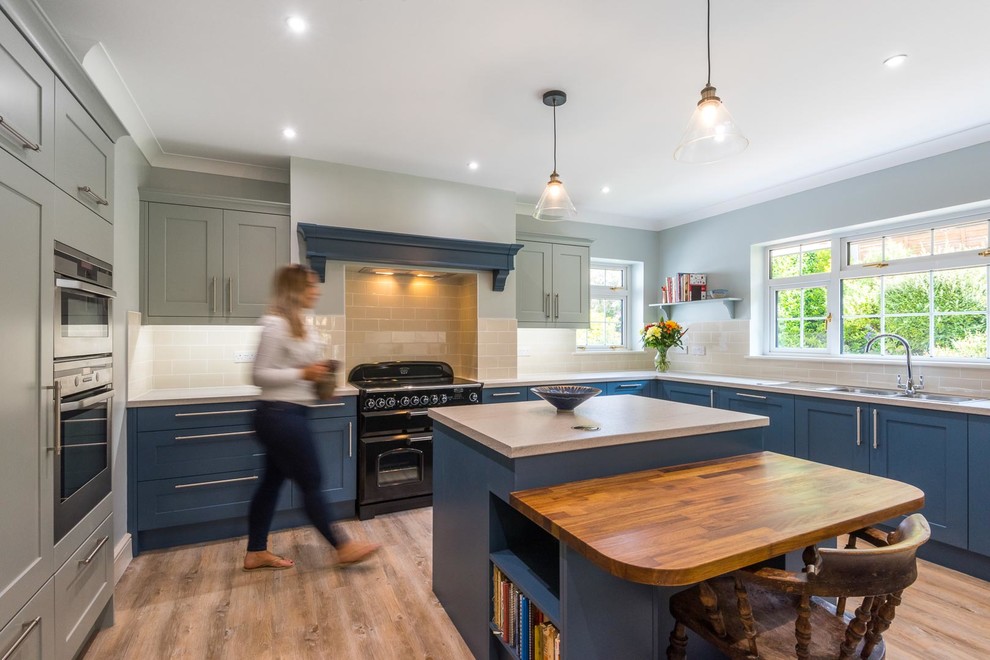 This is an example of a transitional kitchen in Kent with a double-bowl sink, shaker cabinets, blue cabinets, grey splashback, subway tile splashback, black appliances, light hardwood floors, with island and grey benchtop.