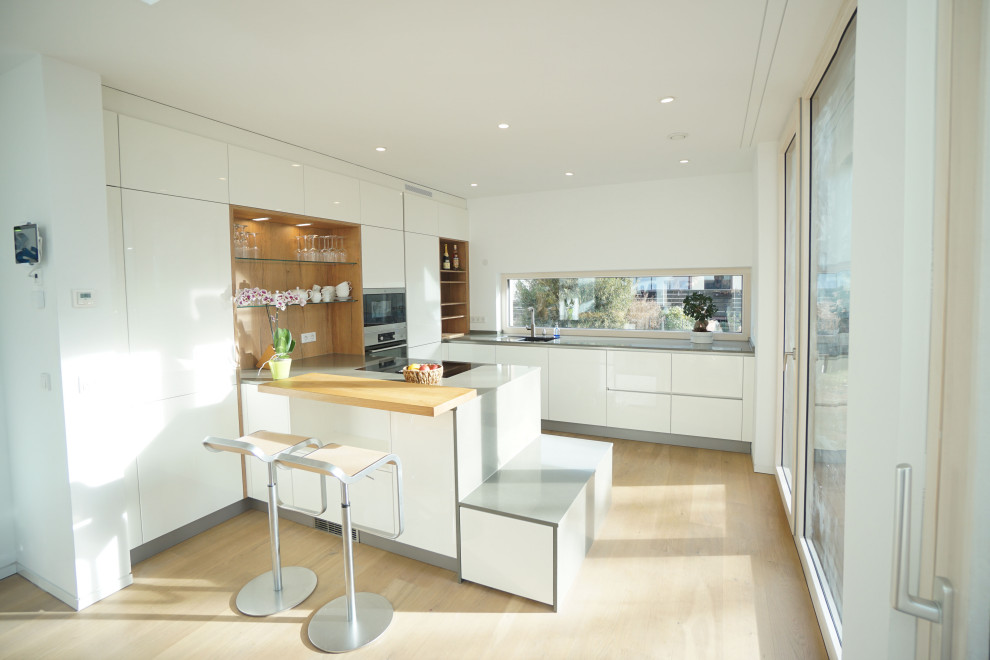 Photo of a mid-sized modern u-shaped open plan kitchen in Munich with an integrated sink, flat-panel cabinets, white cabinets, solid surface benchtops, white splashback, window splashback, stainless steel appliances, light hardwood floors, a peninsula, beige floor and grey benchtop.
