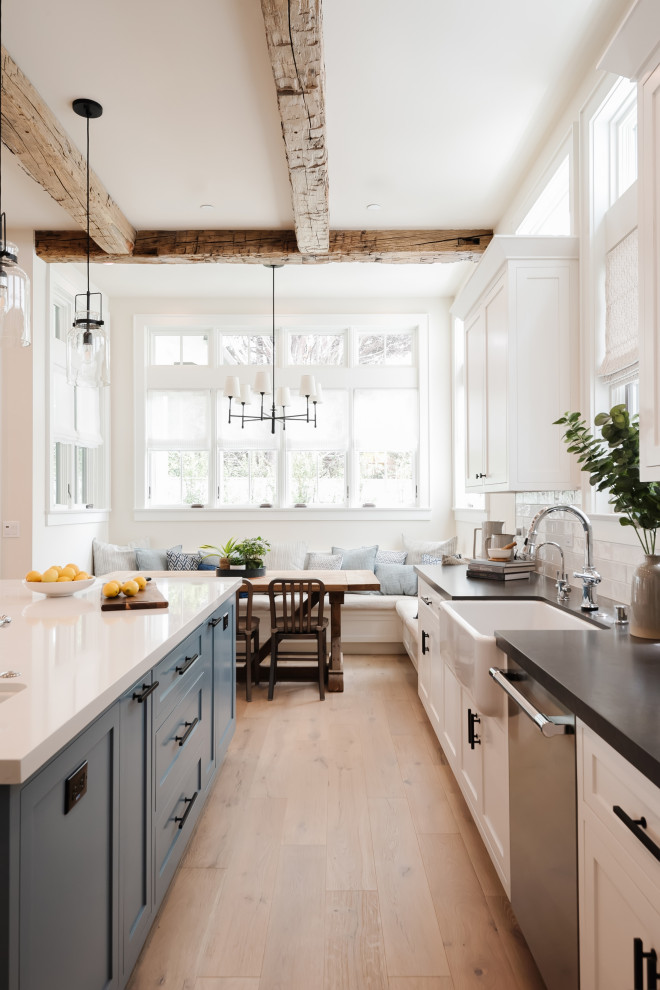 This is an example of a large country u-shaped open plan kitchen in San Diego with a farmhouse sink, shaker cabinets, white cabinets, quartz benchtops, white splashback, porcelain splashback, stainless steel appliances, light hardwood floors, with island, brown floor, black benchtop and exposed beam.