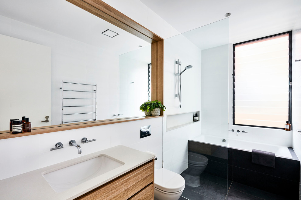 This is an example of a large contemporary master bathroom in Sydney with flat-panel cabinets, a drop-in tub, an open shower, a one-piece toilet, white tile, ceramic tile, white walls, ceramic floors, an undermount sink, engineered quartz benchtops, black floor, an open shower and white benchtops.