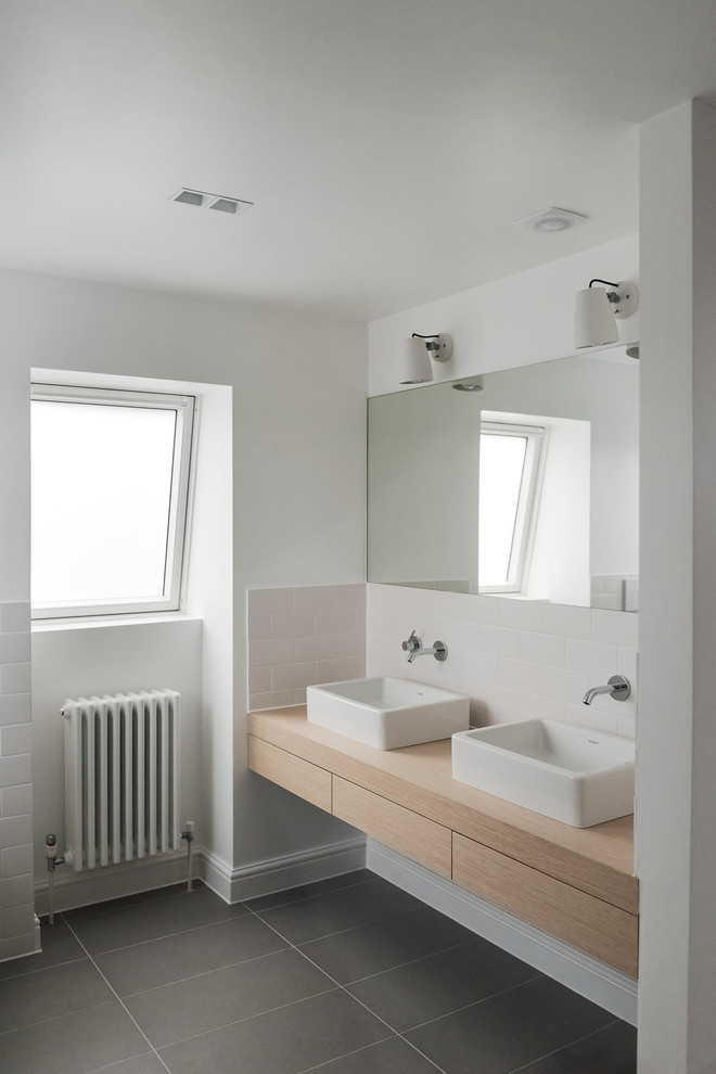 Mid-sized modern kids bathroom in London with flat-panel cabinets, light wood cabinets, a drop-in tub, a shower/bathtub combo, a wall-mount toilet, white tile, subway tile, white walls, cement tiles, a console sink, wood benchtops, grey floor and a hinged shower door.