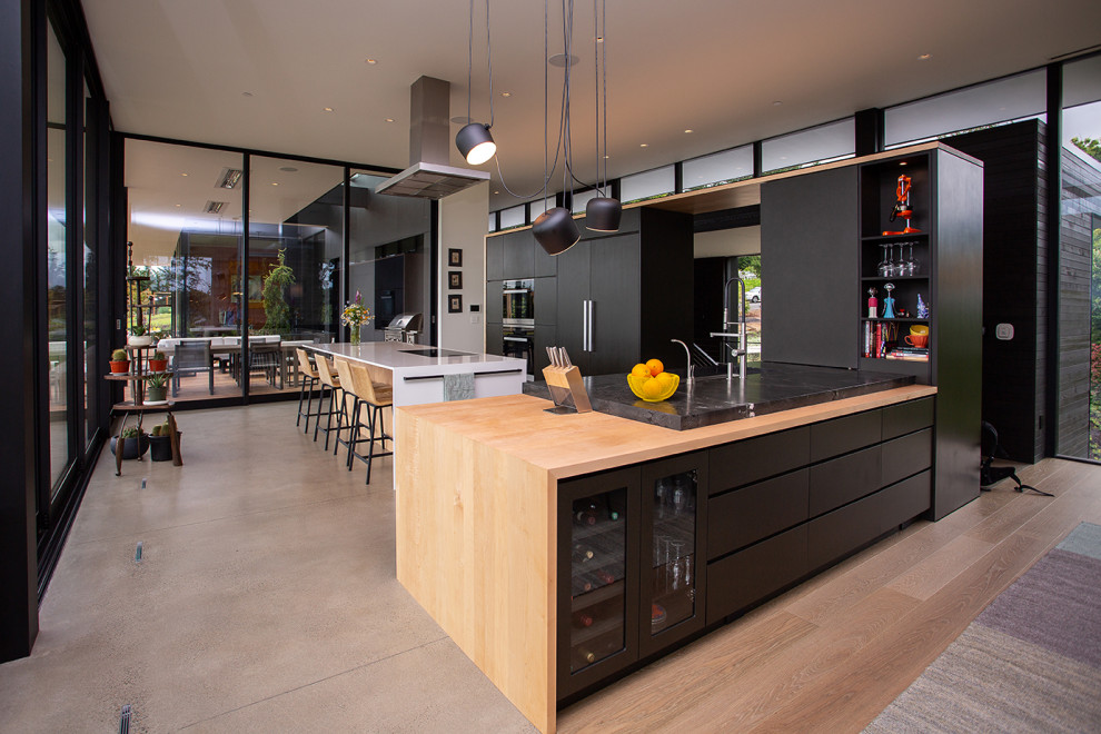 Photo of a modern l-shaped eat-in kitchen in Portland with flat-panel cabinets, black cabinets, wood benchtops, with island, a drop-in sink, panelled appliances, concrete floors and black benchtop.
