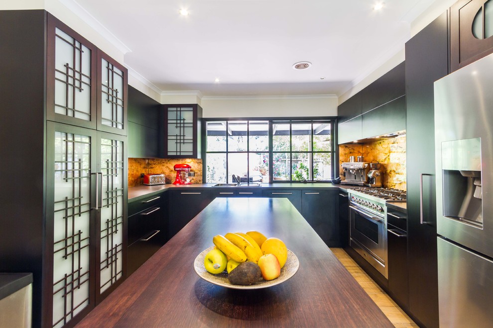 Large asian u-shaped eat-in kitchen in Melbourne with flat-panel cabinets, black cabinets, wood benchtops, metallic splashback, glass sheet splashback, stainless steel appliances, light hardwood floors and with island.