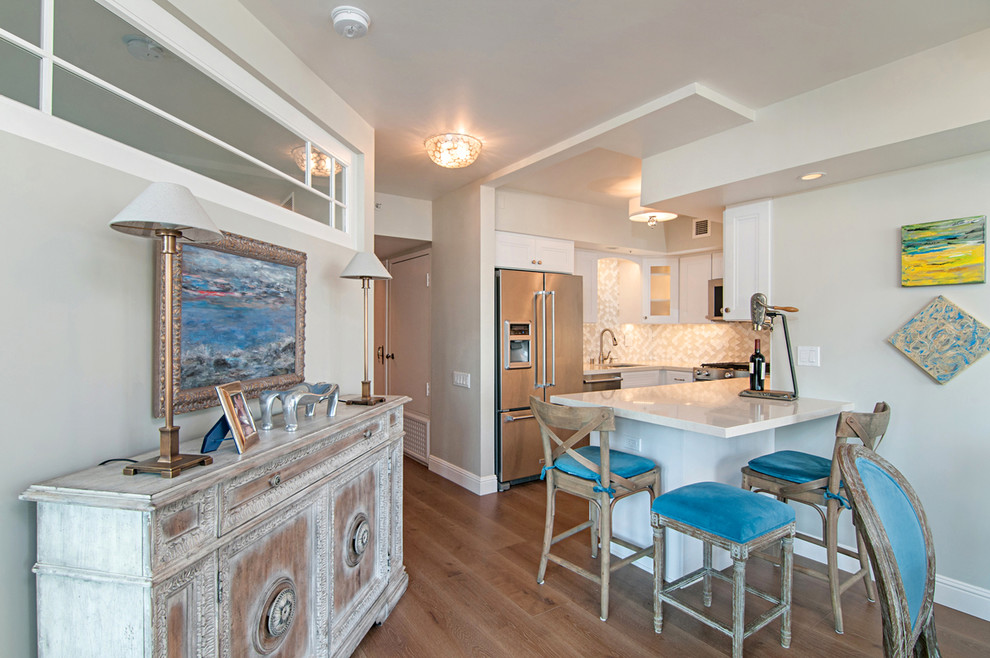 Photo of a small beach style u-shaped open plan kitchen in San Diego with a drop-in sink, white cabinets, white splashback, stainless steel appliances, light hardwood floors and no island.