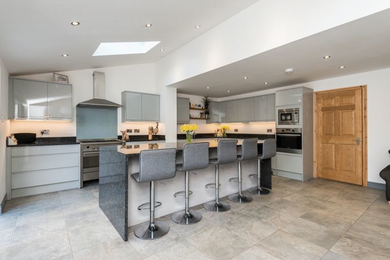 Photo of a large traditional galley eat-in kitchen in London with an integrated sink, flat-panel cabinets, grey cabinets, quartzite benchtops, white splashback, limestone splashback, stainless steel appliances, cement tiles, a peninsula, beige floor and black benchtop.