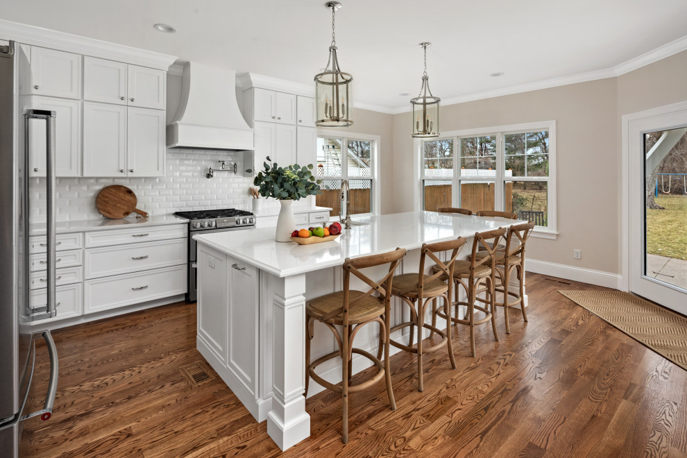 This is an example of a large transitional l-shaped kitchen in St Louis with a farmhouse sink, shaker cabinets, white cabinets, quartzite benchtops, white splashback, subway tile splashback, stainless steel appliances, with island and white benchtop.