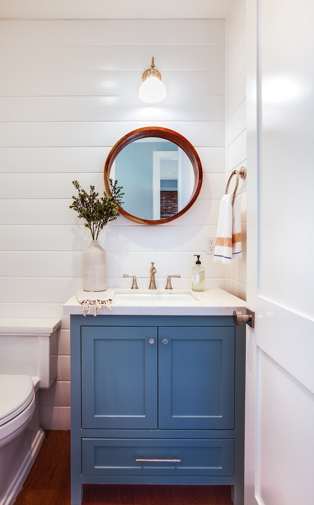 This is an example of a small beach style powder room in Los Angeles with shaker cabinets, blue cabinets, white walls, dark hardwood floors, a wall-mount sink, engineered quartz benchtops, brown floor and white benchtops.